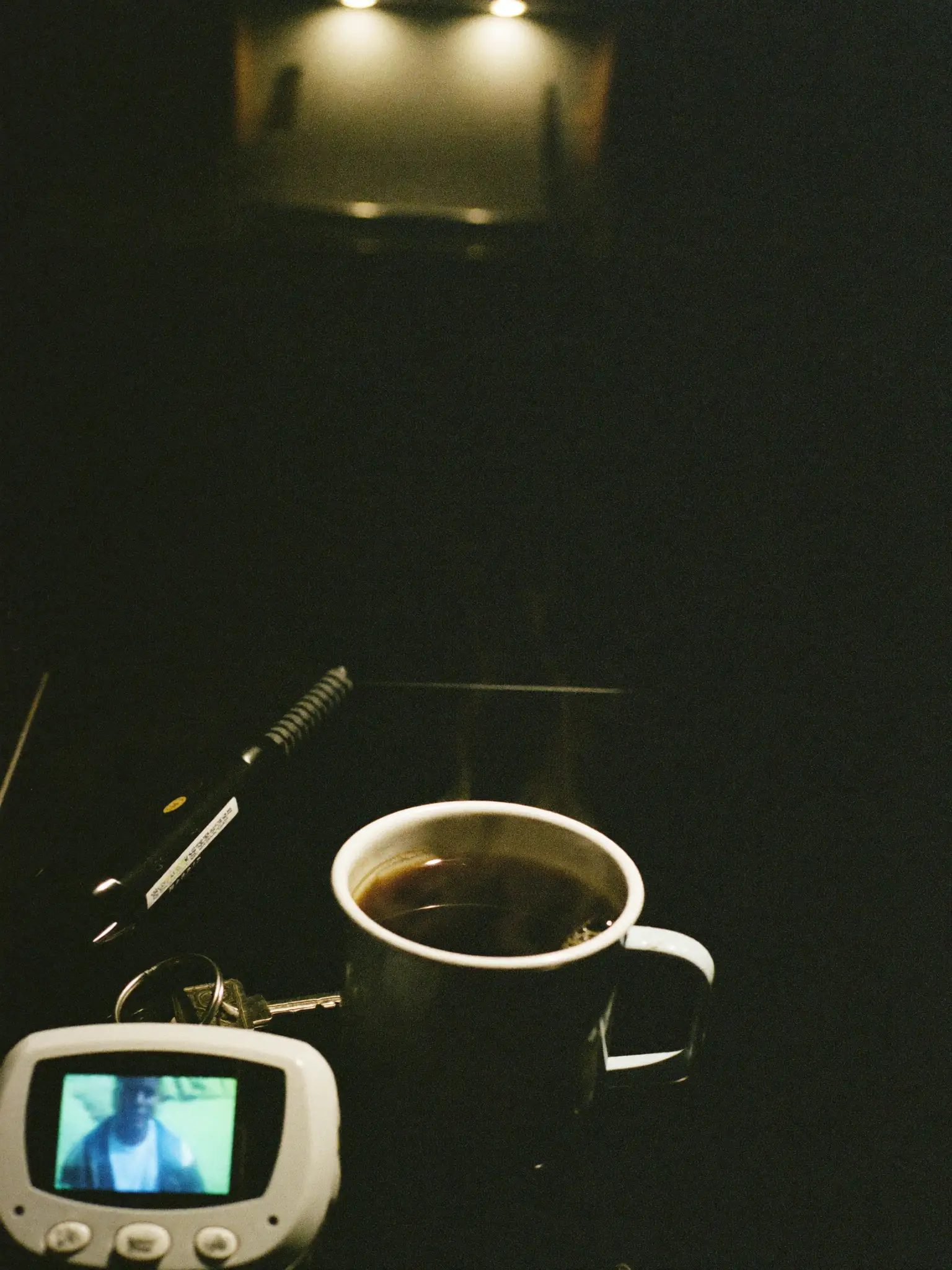 A black mug of coffee next to a baby monitor in a dark kitchen at night.